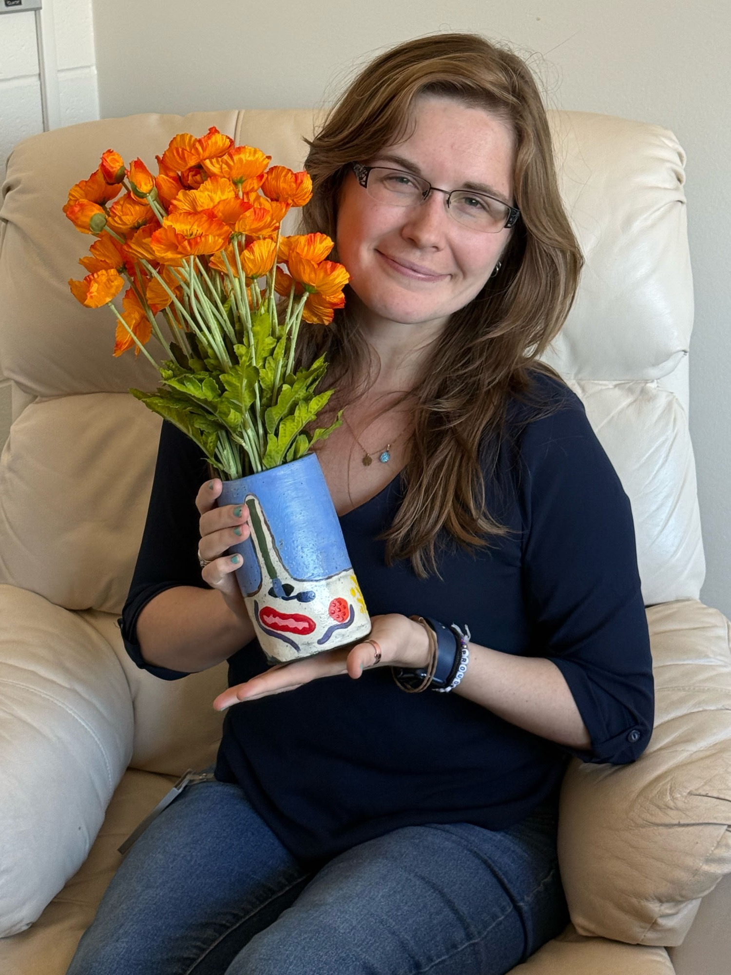 Rachel Turn seated in an armchair, holding a hand-painted vase of orange poppies